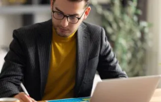 Man working from home office writing down notes on a piece of paper with laptop open