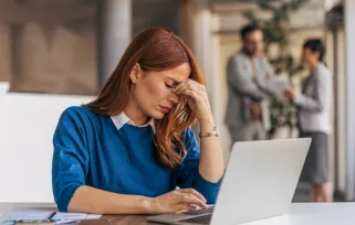 woman in blue sweater sits at her laptop computer at work and is hunched over, appearing stressed or overwhelmed