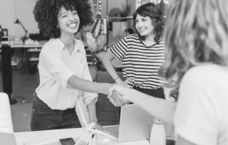 two women shake hands across an interview table while another woman looks on, smiling