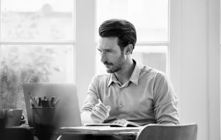 man with beard sitting at laptop with pen and paper