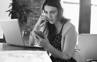 woman sitting at laptop looking nervously at cell phone