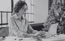 Woman in glasses typing on a laptop with a christmas tree in the background