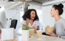 two women sitting at a desk eating lunch and smiling